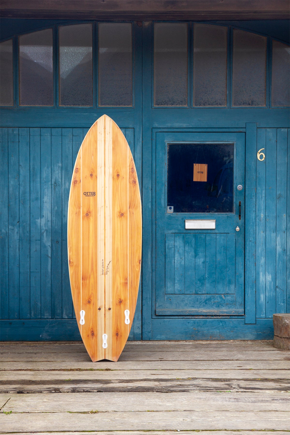 工房の青い扉(No.6)を背景に、美しい節(フシ)のある木目が特徴的な中空構造(ホロー)の木製サーフボード(天然木)のボトム面。埋め込まれた白いフィンプラグと「Otter」の焼印ロゴが、素朴な木の表情の中にモダンな機能性をアクセントとして添える、洗練されたエコラグジュアリーなデザイン。