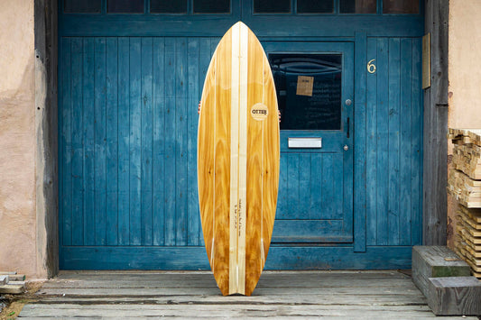 工房の青い扉(No.6)を背景に、ボトム面を披露するフィッシュテールの木製サーフボード。クアッドフィンのセットアップと「Otter」のロゴマークが、クラシックな雰囲気の中でモダンな機能美を主張する、洗練されたエコラグジュアリーなデザイン。