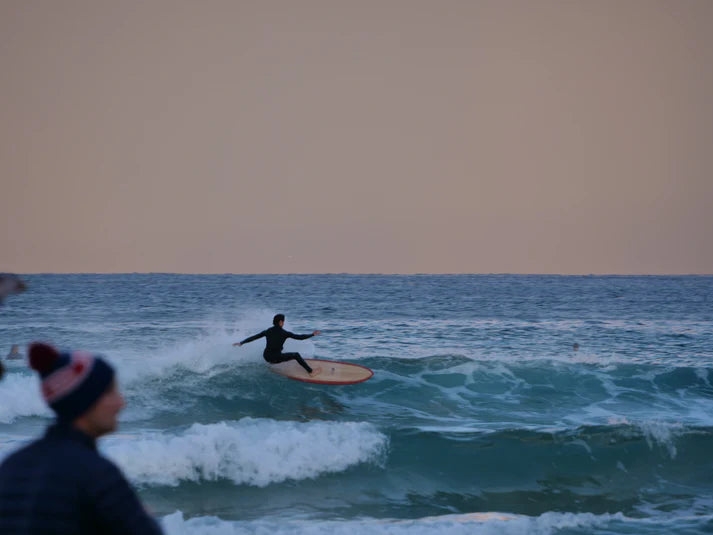 薄明かりの中、波に乗るライダー。遠景のサーファーと天然木のボードが織りなす情景。