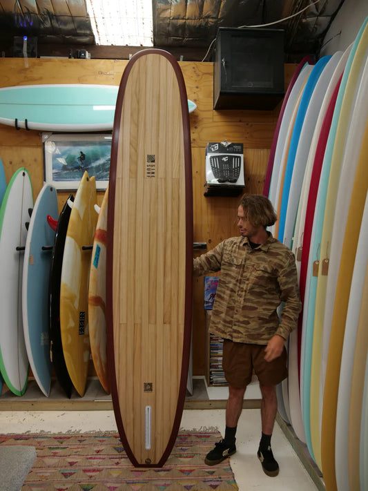 店内で木製ロングボードを立てて展示する男性。美しい木目とワインレッドのレールが印象的なクラシックスタイルのボードが、カラフルなボードに囲まれて際立つ。