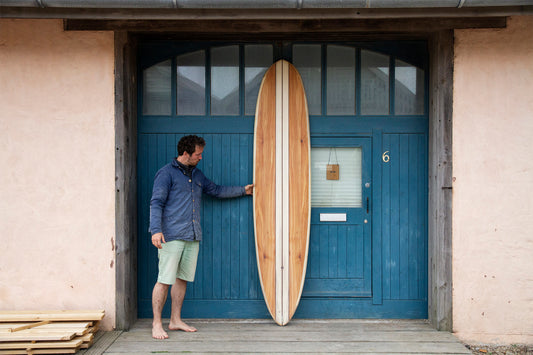 工房の象徴的な青い扉の前で、完成した中空構造(ホロー)の木製サーフボード(天然木)のレールラインを確かめるシェイパー。美しいピンテールのシルエットと洗練されたエコラグジュアリーな佇まい。