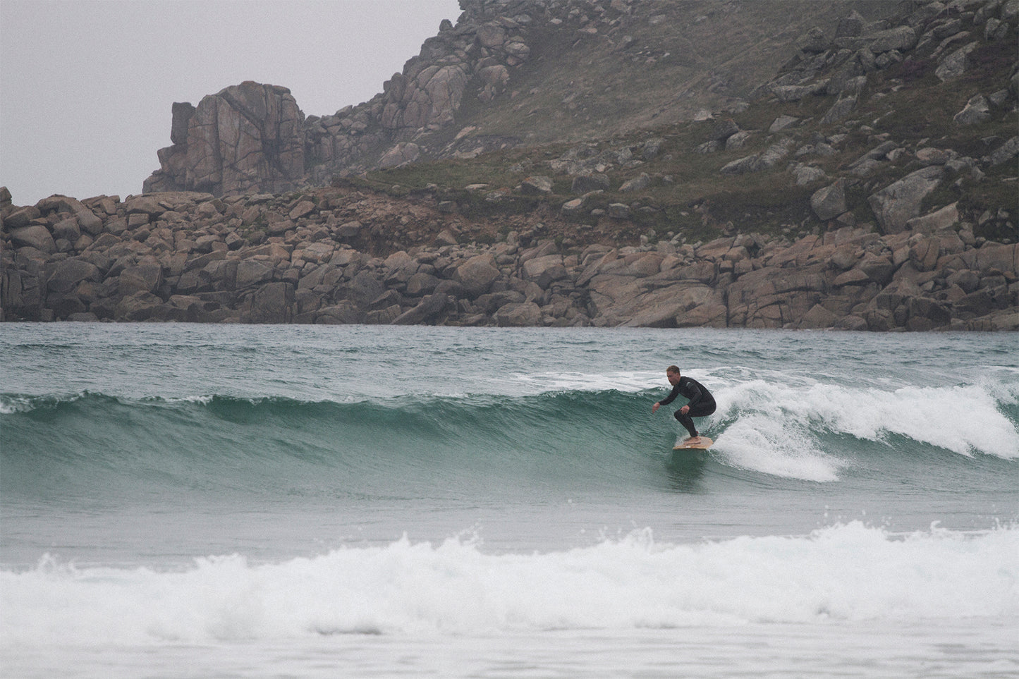 巨岩が転がるワイルドなポイントで、中空構造(ホロー)の木製サーフボード(天然木)を駆るサーファー。自然の厳しさと美しさが同居する場所で波と一体になる、冒険心あふれる大人のサーフスタイル。