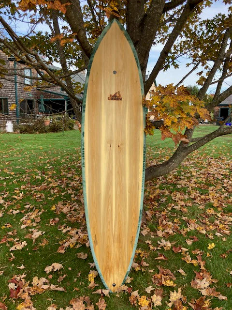 秋の庭に立てかけられた7'4" Tern。シャープなアウトラインと木目が際立つミッドレングス・パフォーマンスボード。