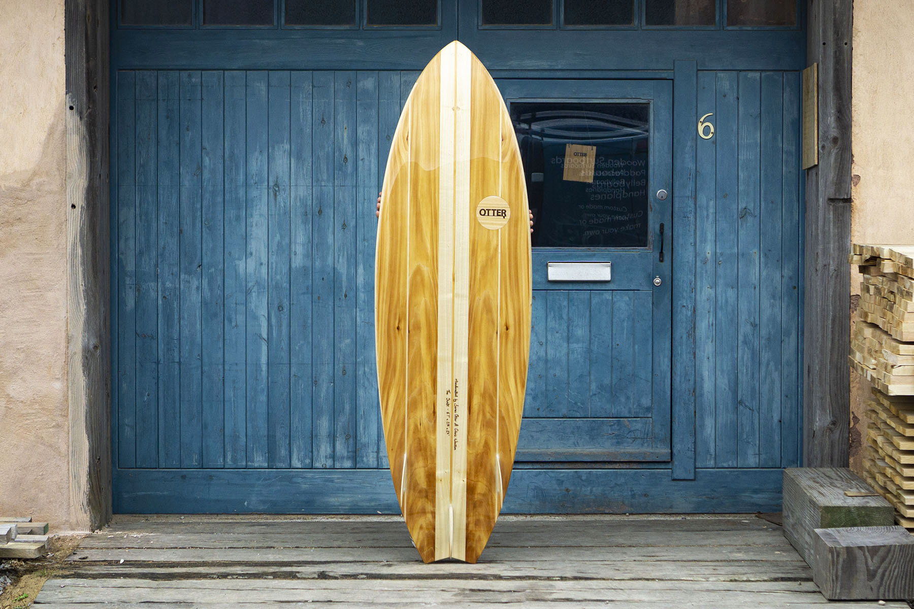 工房の青い扉（No.6）を背景に、ボトム面を披露するフィッシュテールの木製サーフボード。クアッドフィンのセットアップと「Otter」のロゴマークが、クラシックな雰囲気の中でモダンな機能美を主張する、洗練されたエコラグジュアリーなデザイン。
