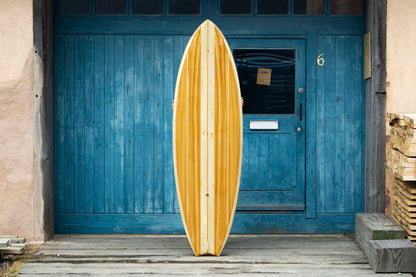 作り手の手に支えられ、誇らしげに掲げられたフィッシュボード。左右対称に美しく配置された木材と、複雑な曲線を描くテールの処理は、中空構造（ホロー）の木製サーフボード（天然木）が単なる道具ではなく、上質なクラフトマンシップによる工芸品であることを証明している。