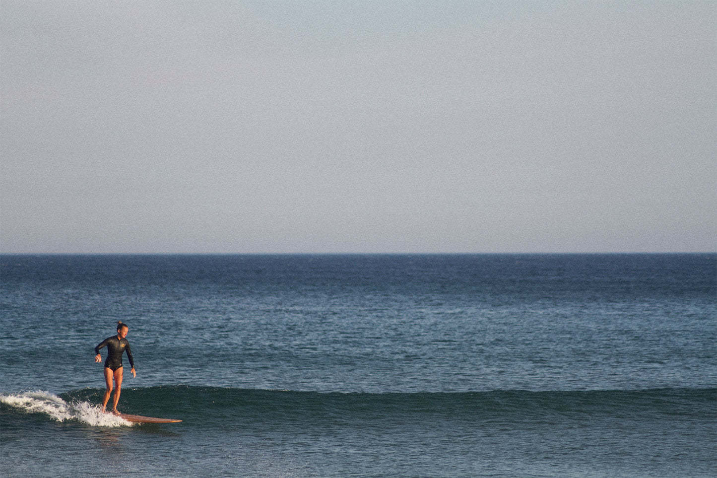 水平線に溶け込むようなブルーの海で、中空構造（ホロー）の木製サーフボード（天然木）の上を優雅に歩くサーファー。ミニマルな美しさとクラシックなスタイルが融合した、洗練されたエコラグジュアリーな一コマ。