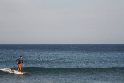 水平線に溶け込むようなブルーの海で、中空構造（ホロー）の木製サーフボード（天然木）の上を優雅に歩くサーファー。ミニマルな美しさとクラシックなスタイルが融合した、洗練されたエコラグジュアリーな一コマ。