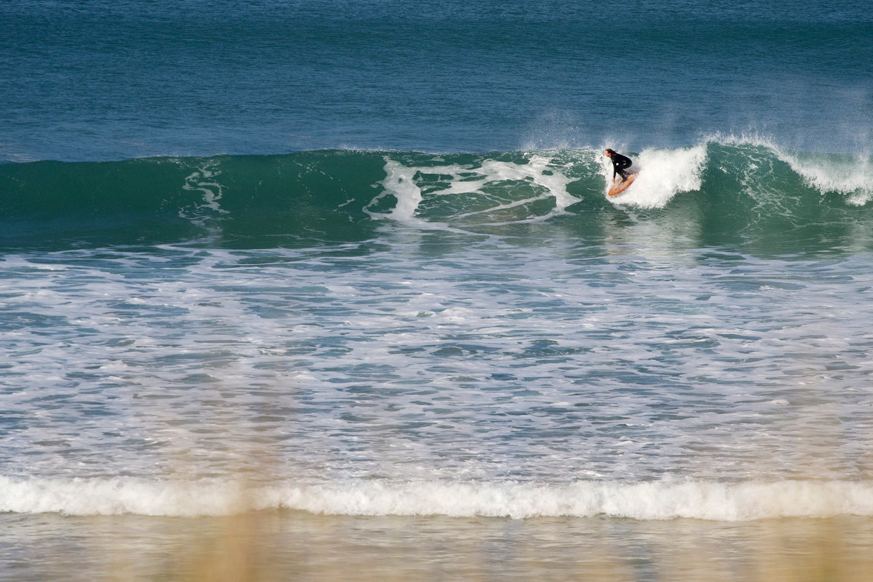 鮮やかなブルーの海と、波のトップでターンを描くサーファー。白いスープと深い青のコントラストの中で、中空構造（ホロー）の木製サーフボード（天然木）の暖色が鮮明に浮かび上がる、洗練されたエコラグジュアリーなアクションシーン。