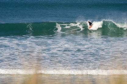 鮮やかなブルーの海と、波のトップでターンを描くサーファー。白いスープと深い青のコントラストの中で、中空構造（ホロー）の木製サーフボード（天然木）の暖色が鮮明に浮かび上がる、洗練されたエコラグジュアリーなアクションシーン。