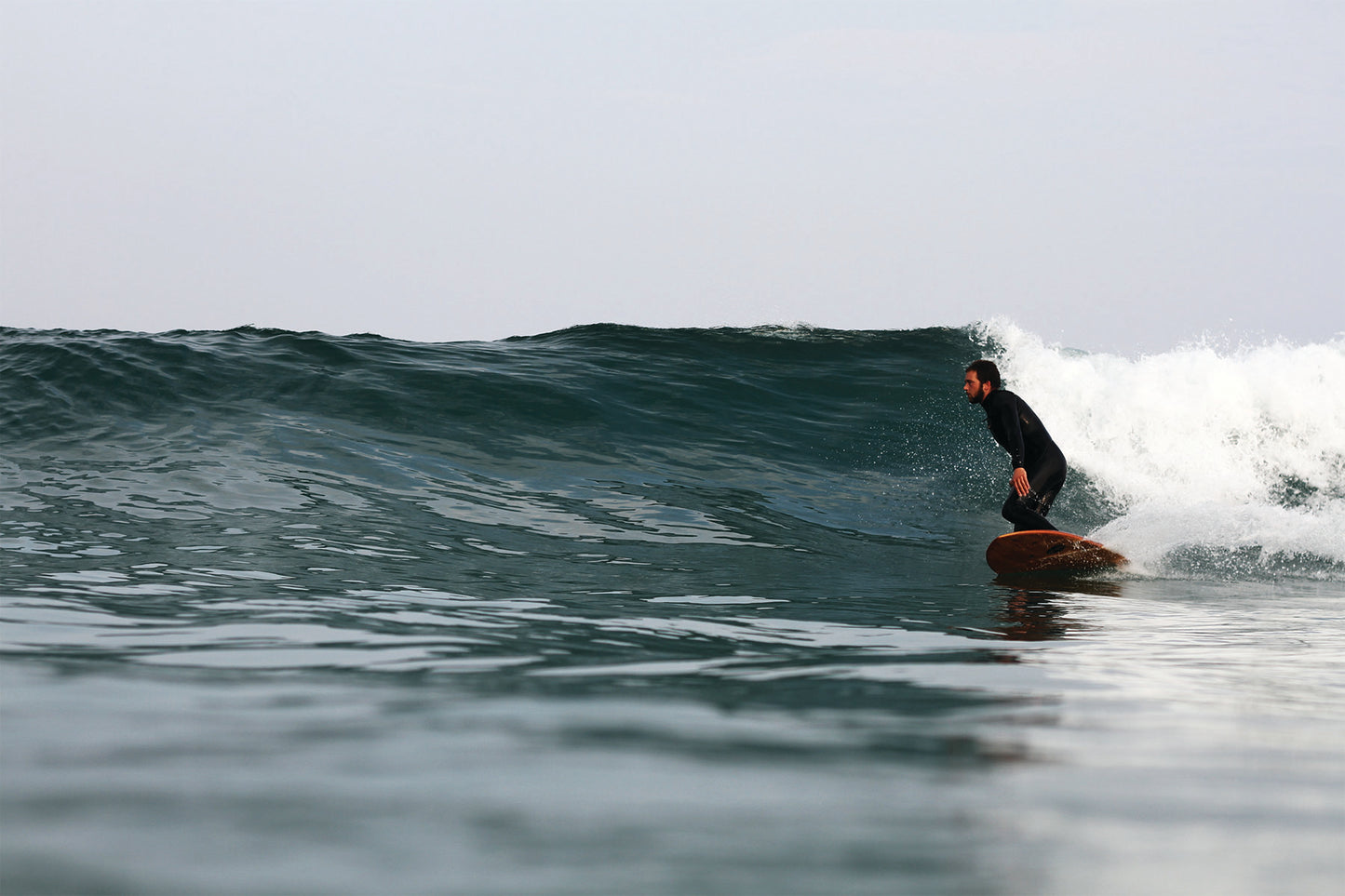 滑らかな水面（グラッシー）と深いグリーンの波のフェイスを、中空構造（ホロー）の木製サーフボード（天然木）が静かに切り裂く。水面ギリギリのローアングルから捉えたショットは、ボードの優美なアウトラインと、波と一体化する洗練されたエコラグジュアリーなライディングを強調する。