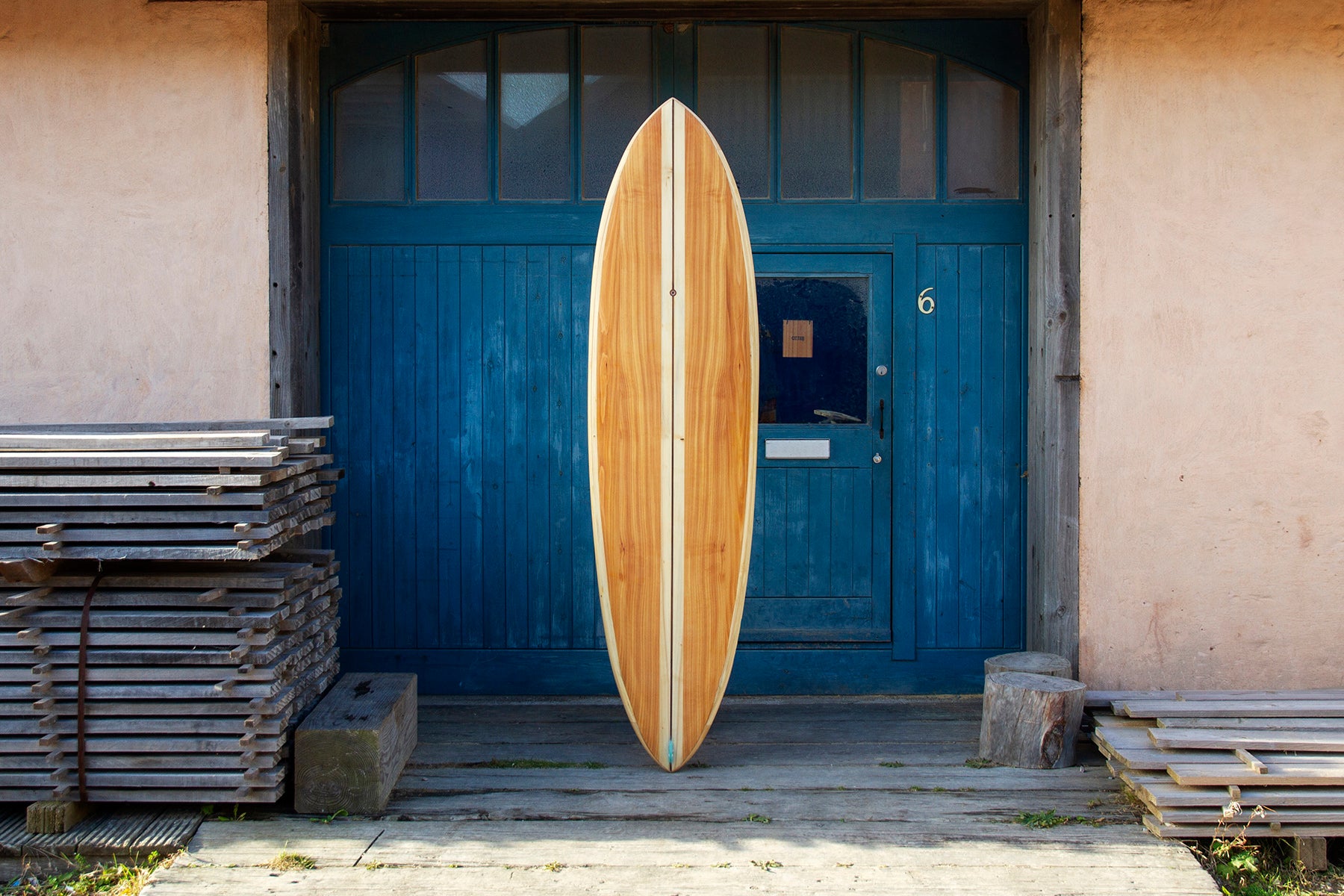 工房の青い扉を背景に、静かな存在感を放つ中空構造（ホロー）の木製サーフボード（天然木）。中央を貫く太めのストリンガーがモダンな印象を与える、大人の遊び心を凝縮したエコラグジュアリーな一本。
