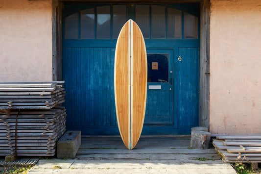 工房の青い扉を背景に、静かな存在感を放つ中空構造（ホロー）の木製サーフボード（天然木）。中央を貫く太めのストリンガーがモダンな印象を与える、大人の遊び心を凝縮したエコラグジュアリーな一本。
