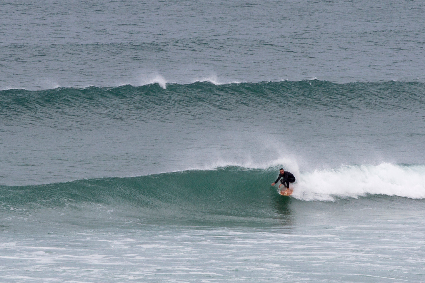 幾重にも重なるうねりの中、クリーンなフェイスでボトムターンを決めるサーファーと中空構造（ホロー）の木製サーフボード（天然木）。エメラルドグリーンの波の壁と、ボードのナチュラルな色彩が美しいコントラストを描く、洗練されたエコラグジュアリーなライディング。