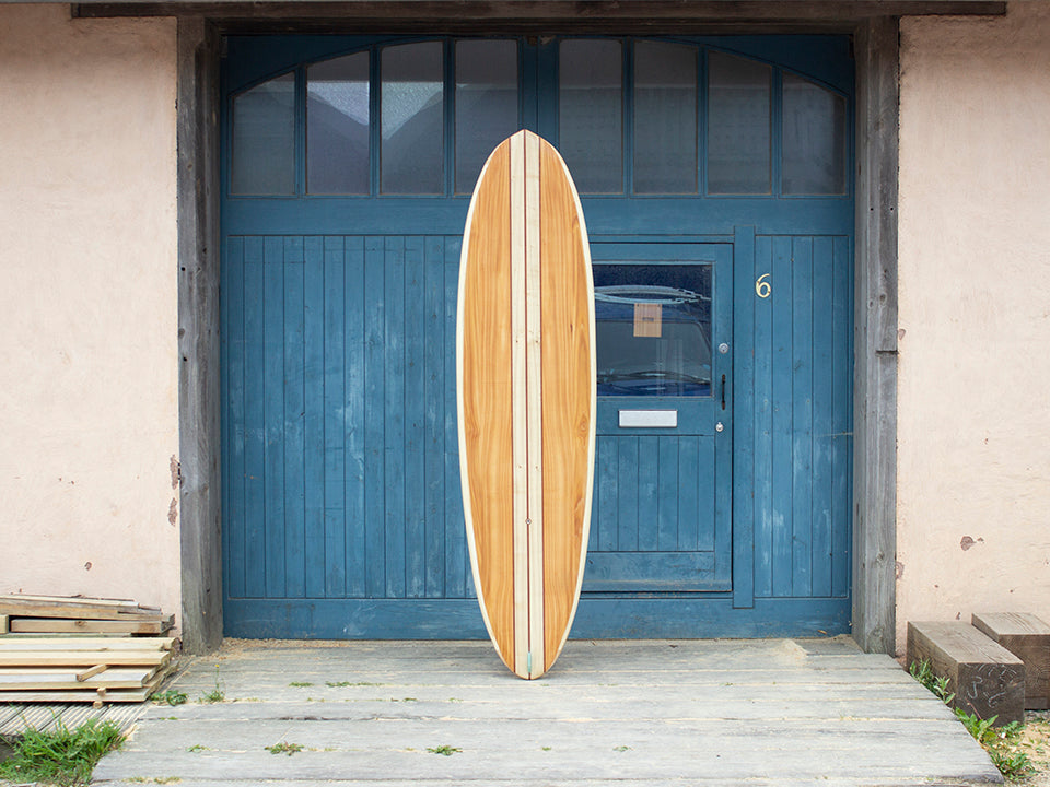 生まれた場所である工房の前で、その完成度を誇るかのような中空構造（ホロー）の木製サーフボード（天然木）。職人の手仕事によって張り合わされた木々の調和と、滑らかなノーズからテールへの曲線美が宿る上質なクラフトマンシップ。