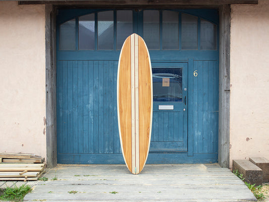 生まれた場所である工房の前で、その完成度を誇るかのような中空構造（ホロー）の木製サーフボード（天然木）。職人の手仕事によって張り合わされた木々の調和と、滑らかなノーズからテールへの曲線美が宿る上質なクラフトマンシップ。