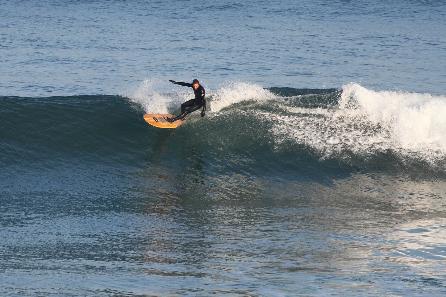 深いブルーの波と、中空構造（ホロー）の木製サーフボード（天然木）の黄金色のコントラスト。スプレーを上げながら描くアグレッシブなターン、その瞬間に垣間見える「Otter」ロゴと洗練されたエコラグジュアリーなデザイン。