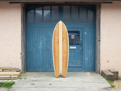 工房のアイコニックな青い扉（No.6）の前に直立する、フィッシュテールの中空構造（ホロー）の木製サーフボード（天然木）。中央の明るいストリンガーと両サイドの暖色系ウッドのコントラストが、ヴィンテージ感漂うブルーの背景に映える、洗練されたエコラグジュアリーなポートレート。