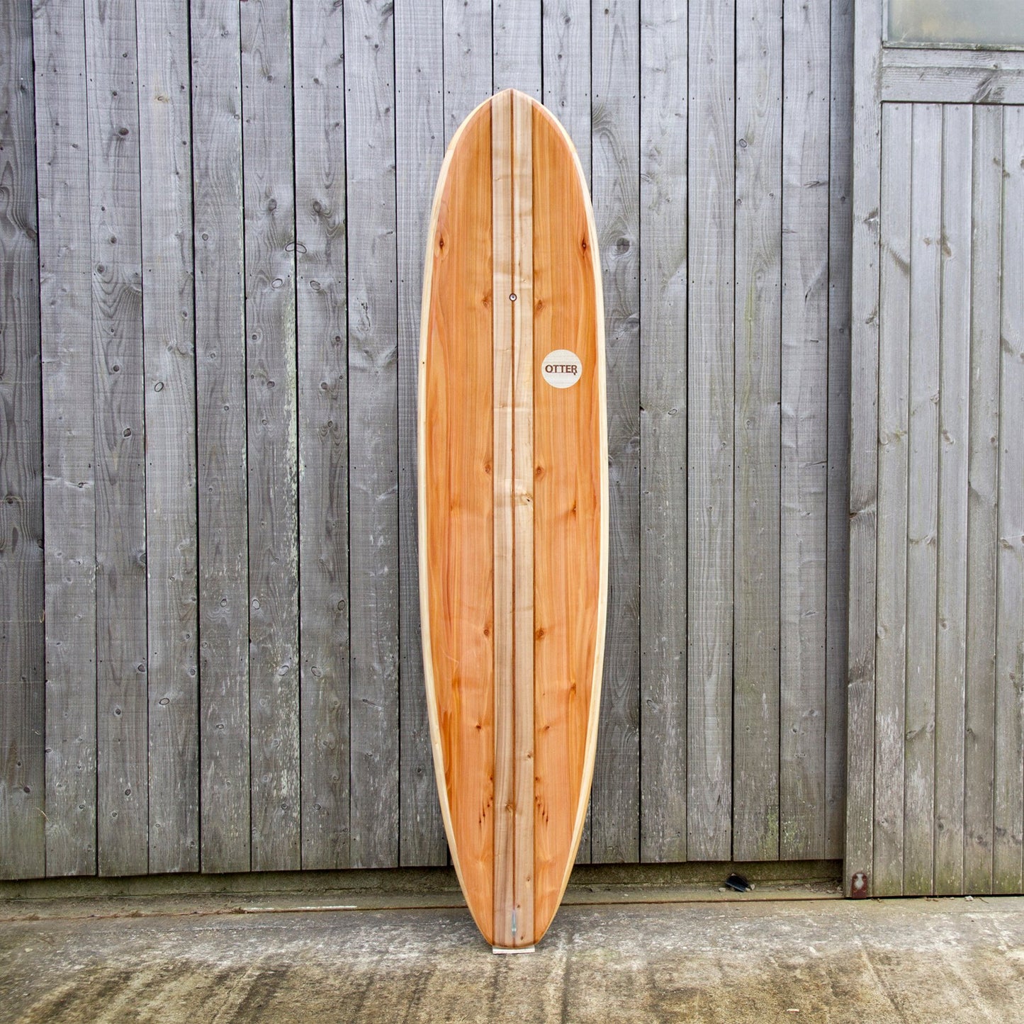 経年変化した壁面と、真新しい中空構造（ホロー）の木製サーフボード（天然木）の対比。豊かな木目と節（フシ）の表情が、飾れる・乗れるサステナブルな機能美を主張する。