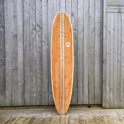 経年変化した壁面と、真新しい中空構造（ホロー）の木製サーフボード（天然木）の対比。豊かな木目と節（フシ）の表情が、飾れる・乗れるサステナブルな機能美を主張する。