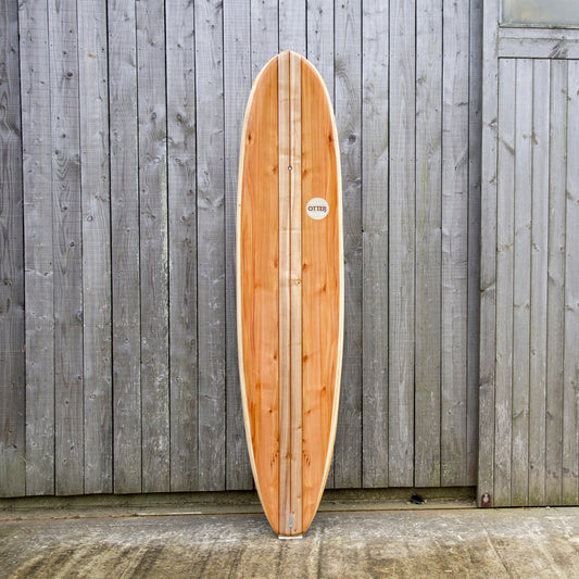 経年変化した壁面と、真新しい中空構造（ホロー）の木製サーフボード（天然木）の対比。豊かな木目と節（フシ）の表情が、飾れる・乗れるサステナブルな機能美を主張する。