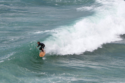 白波（スープ）が渦巻くセクションを抜けようとするサーファーと、中空構造（ホロー）の木製サーフボード（天然木）。俯瞰で捉えた構図が、荒れた水面でも安定感を失わないボードの優れた設計と洗練されたエコラグジュアリーな機能美を証明する。