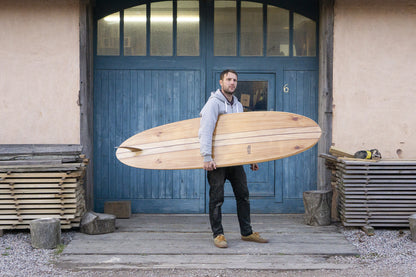 工房の象徴的な青い扉の前で、中空構造（ホロー）の木製サーフボード（天然木）を横に掲げる男性。水平に保持されたボードの美しいアウトラインとダークカラーのストリンガー、中央のOtterロゴが視線を惹きつける、洗練されたエコラグジュアリーなポートレート。