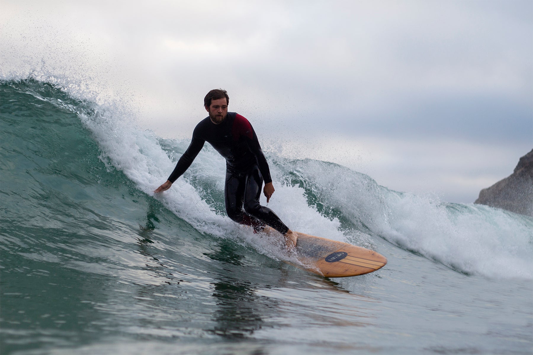 赤いショルダーパッチのウェットスーツが映える、中空構造（ホロー）の木製サーフボード（天然木）でのクルージング。自然のリズムに身を委ねる、集中と解放が共存する大人のサーフシーン。