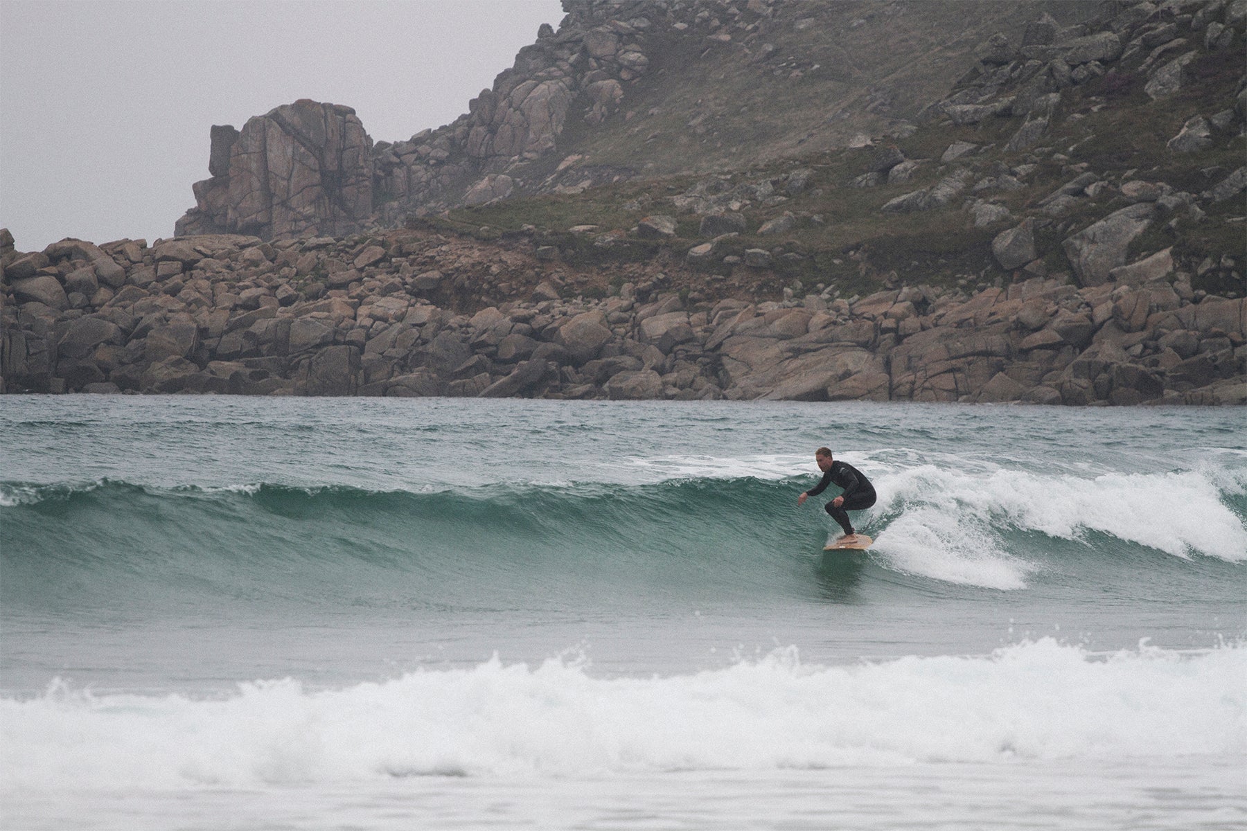 巨岩が転がるワイルドなポイントで、中空構造（ホロー）の木製サーフボード（天然木）を駆るサーファー。自然の厳しさと美しさが同居する場所で波と一体になる、冒険心あふれる大人のサーフスタイル。