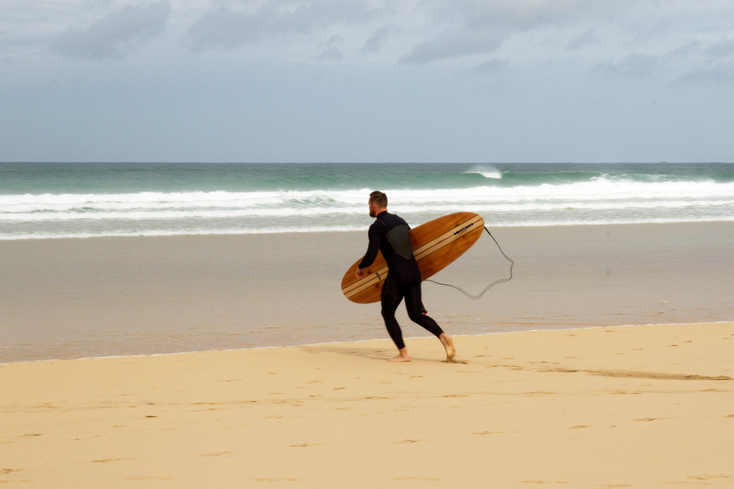 目の前のブレイクに心を躍らせ、小走りで海へ向かう瞬間。中空構造（ホロー）の木製サーフボード（天然木）と共に過ごす、純粋な喜びと冒険心に満ちた大人のサーフスタイル。