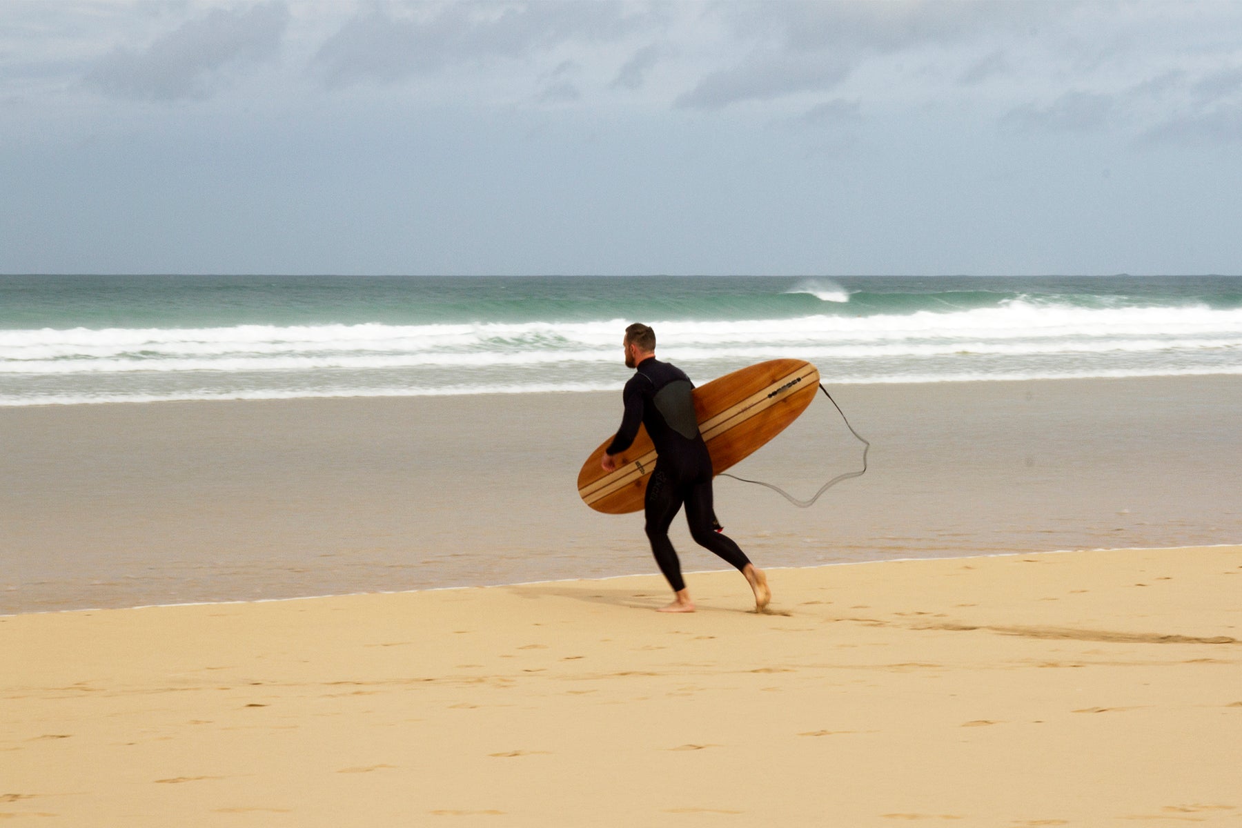 目の前のブレイクに心を躍らせ、小走りで海へ向かう瞬間。中空構造（ホロー）の木製サーフボード（天然木）と共に過ごす、純粋な喜びと冒険心に満ちた大人のサーフスタイル。