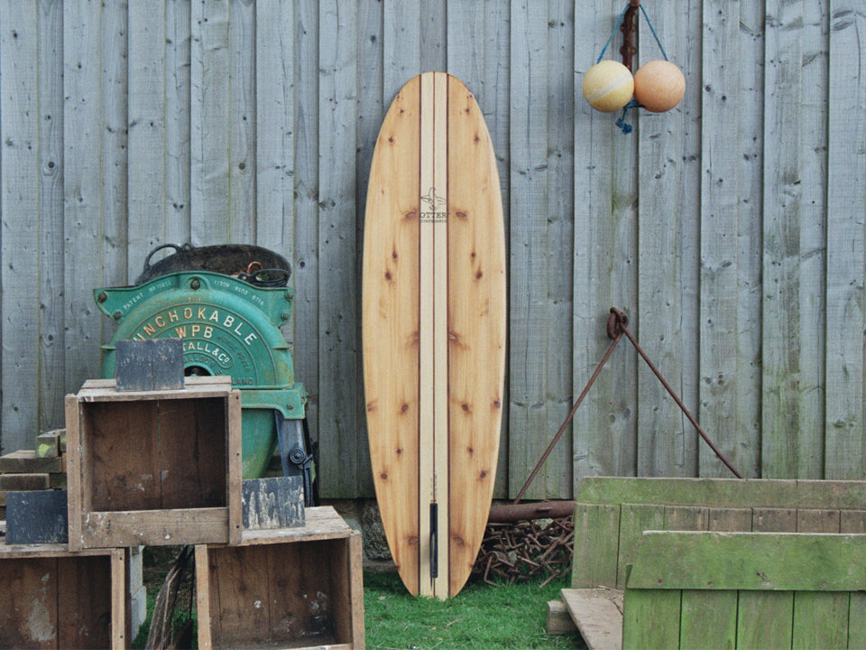 風化した木の板壁、錆びた鉄、そして磨き上げられた中空構造（ホロー）の木製サーフボード（天然木）の対比。吊るされたブイや木箱の質感が、ボードの滑らかで有機的な美しさを際立たせる、飾れる・乗れるサステナブルな機能美。