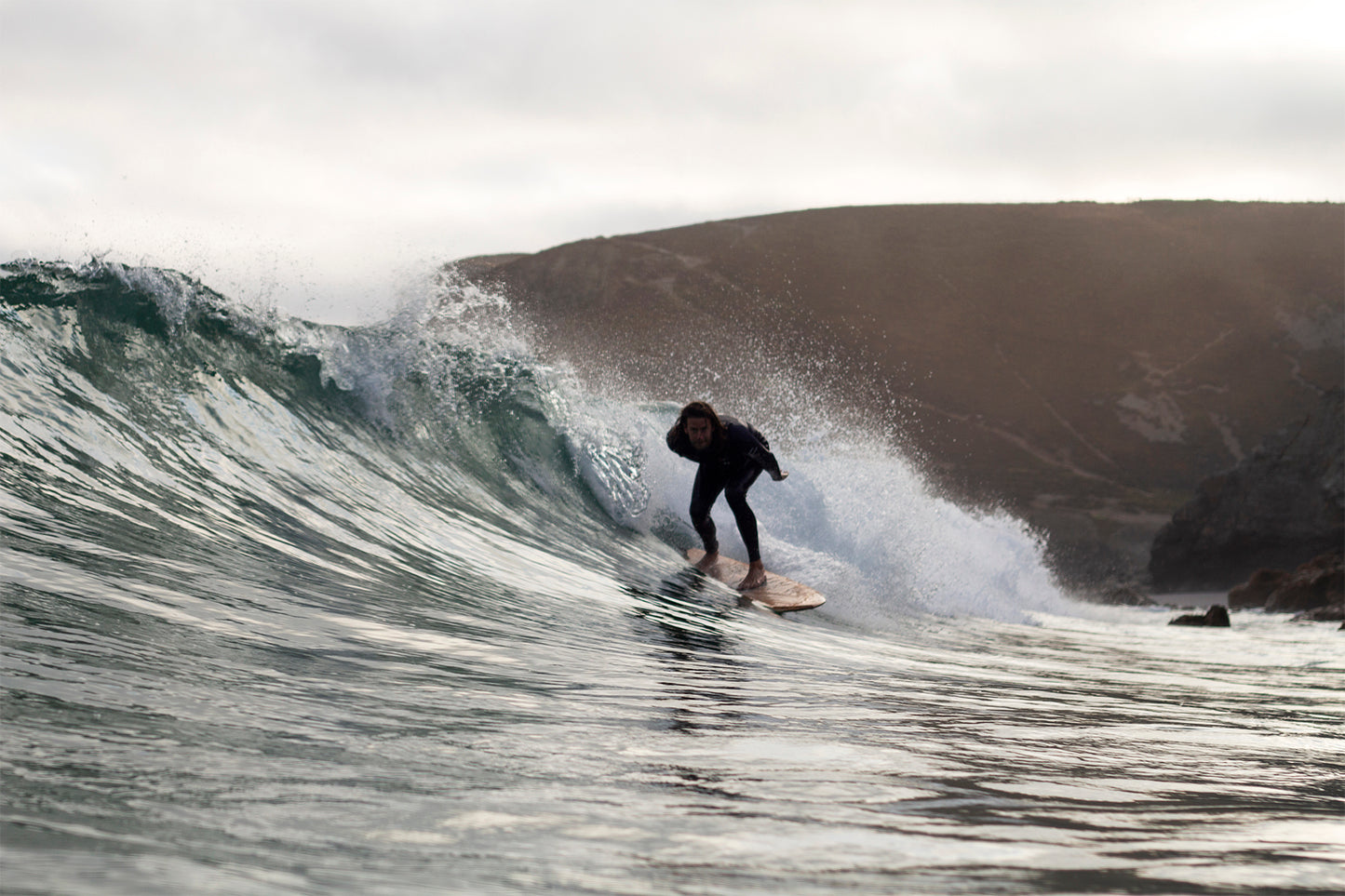 荒涼とした崖を背景に、大きなスプレーを上げて波を刻むサーファーと中空構造（ホロー）の木製サーフボード（天然木）。ダイナミックなアクションと静寂な風景が交差する瞬間、ボードの有機的なフォルムが洗練されたエコラグジュアリーな存在感を放つ。