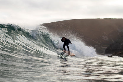 荒涼とした崖を背景に、大きなスプレーを上げて波を刻むサーファーと中空構造（ホロー）の木製サーフボード（天然木）。ダイナミックなアクションと静寂な風景が交差する瞬間、ボードの有機的なフォルムが洗練されたエコラグジュアリーな存在感を放つ。