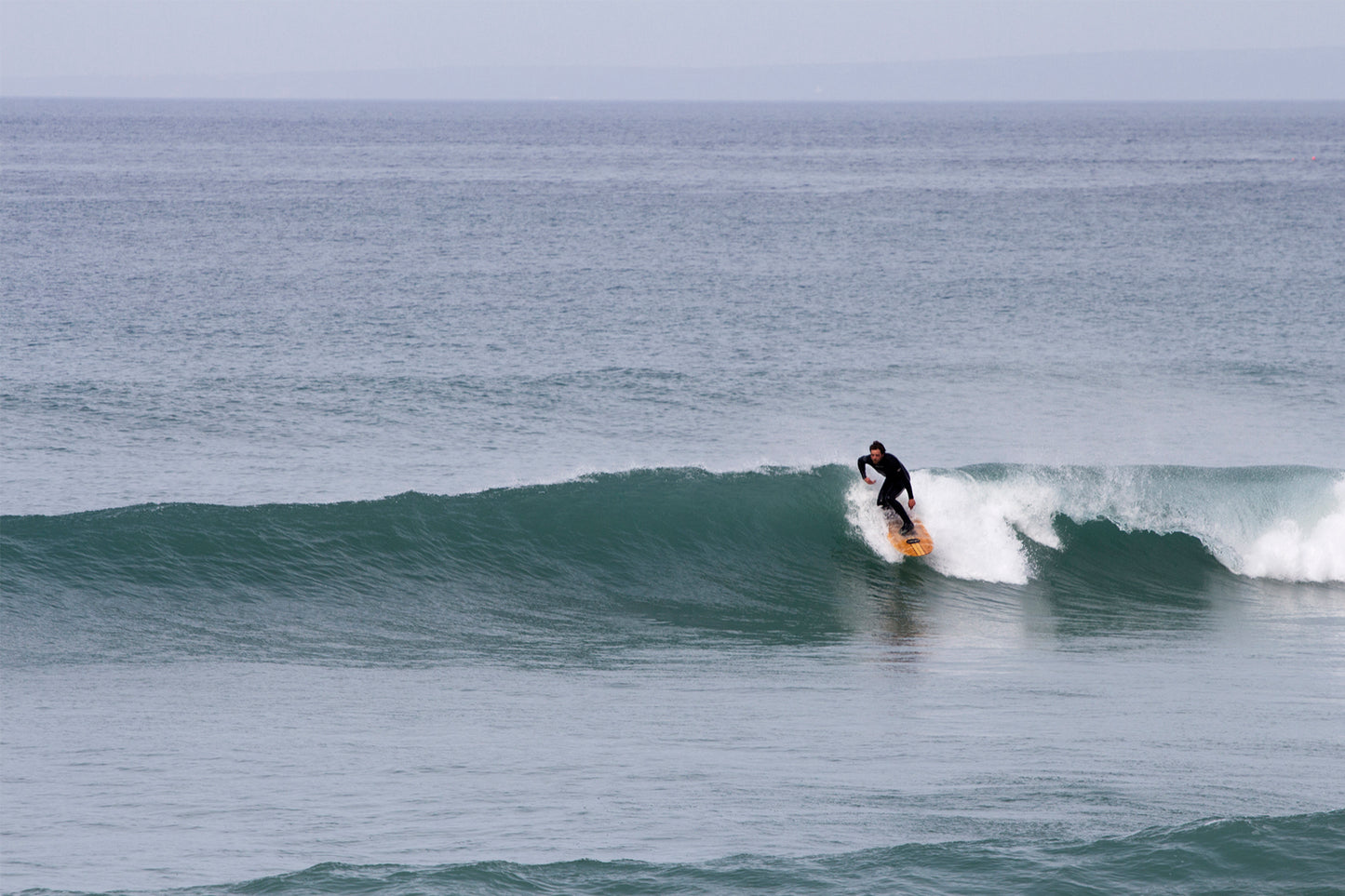 広い水平線を背景に、緑青色の波をトリムするサーファーと中空構造（ホロー）の木製サーフボード（天然木）。波のフェイスを滑るボードの軌跡が、洗練されたエコラグジュアリーなライディングを際立たせる一枚。