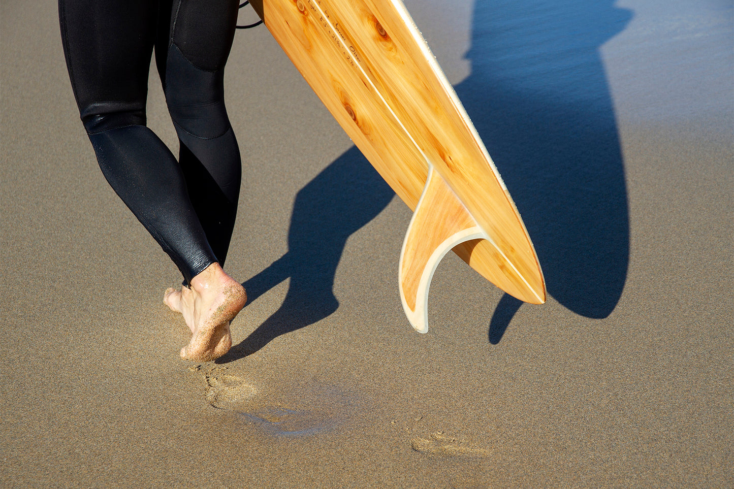 波乗りを終えた充実感か、これからの期待か。中空構造（ホロー）の木製サーフボード（天然木）を抱えて歩くビーチサイド。大地と繋がり、自然のリズムを感じる大人のサーフスタイル。