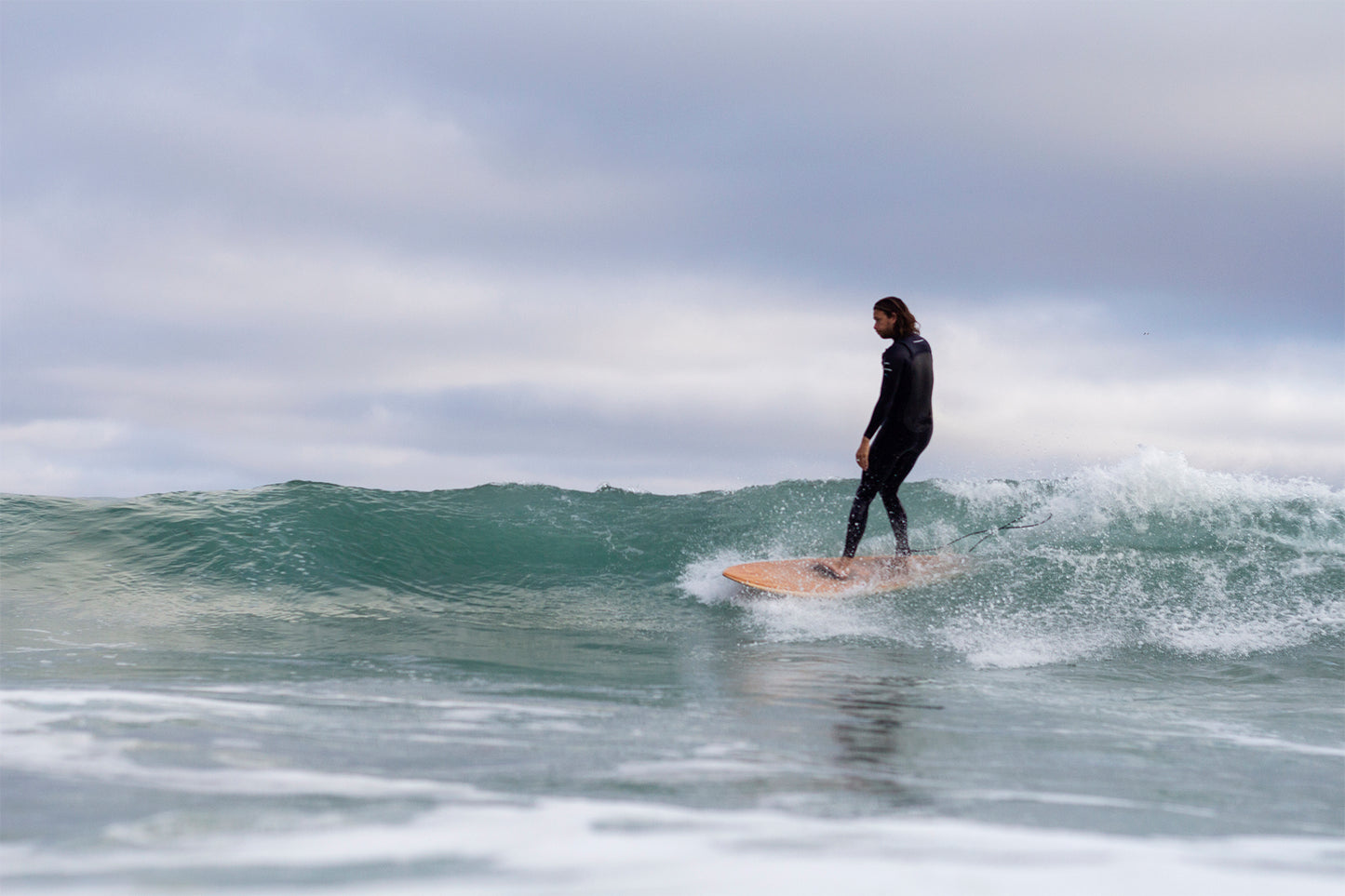 曇り空の下、優雅にクロスステップを踏むサーファーと中空構造（ホロー）の木製サーフボード（天然木）。クラシックなロングボードスタイルと、ボードの安定感が織りなす、洗練されたエコラグジュアリーなライディングシーン。
