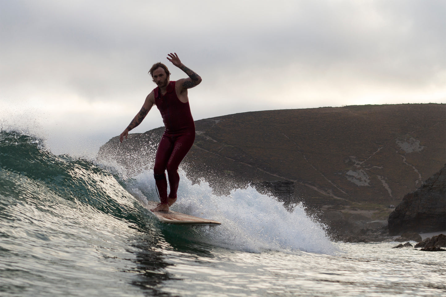 荒涼とした崖を背景に、中空構造（ホロー）の木製サーフボード（天然木）を操る赤いウェットスーツのサーファー。自然の厳しさとウッドの温もりが同居する、洗練されたエコラグジュアリーなアクションシーン。