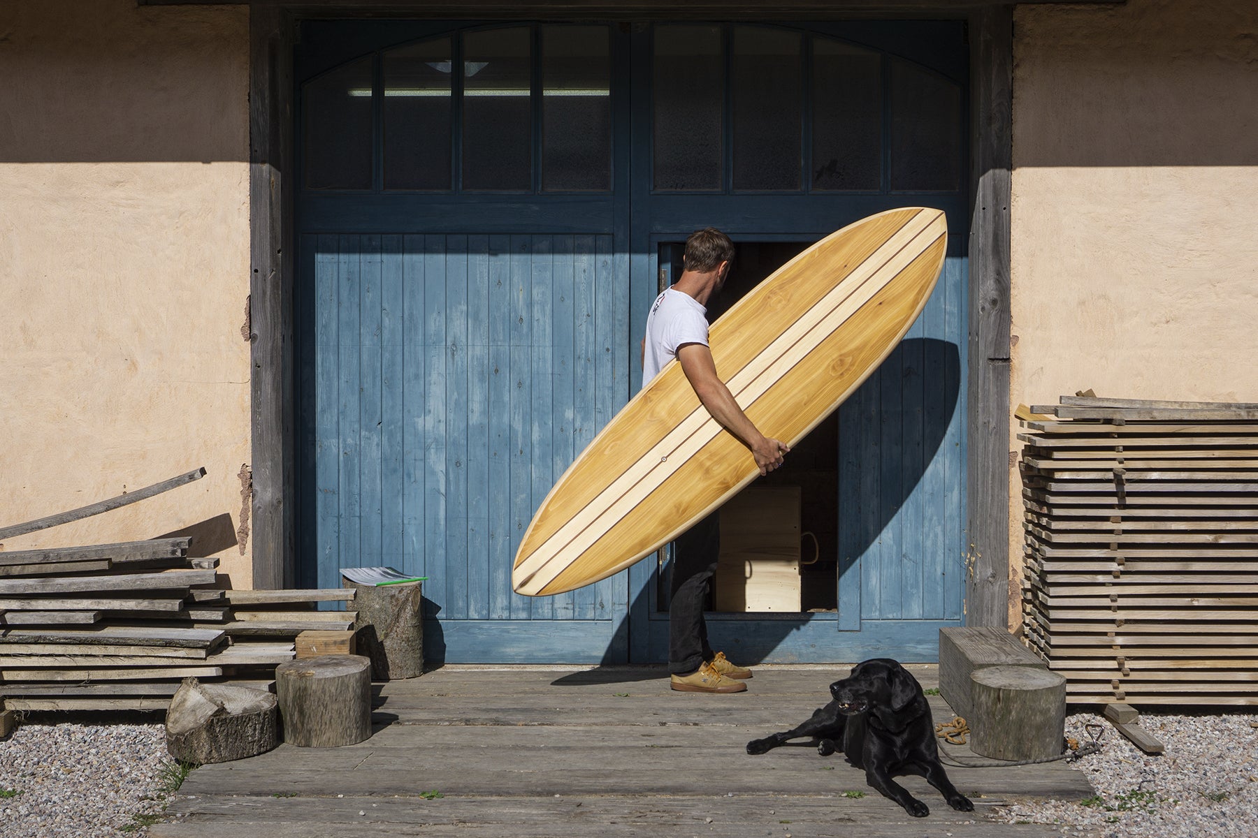 制作の現場である工房へ、中空構造（ホロー）の木製サーフボード（天然木）を運び込む一コマ。職人の手仕事と愛犬との時間が織りなす、温もり溢れる上質なウッドのある暮らし。
