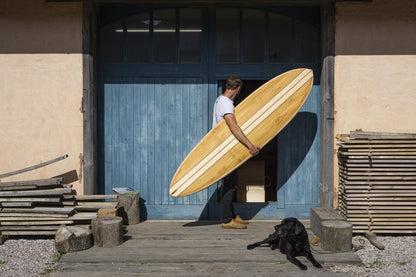 制作の現場である工房へ、中空構造（ホロー）の木製サーフボード（天然木）を運び込む一コマ。職人の手仕事と愛犬との時間が織りなす、温もり溢れる上質なウッドのある暮らし。