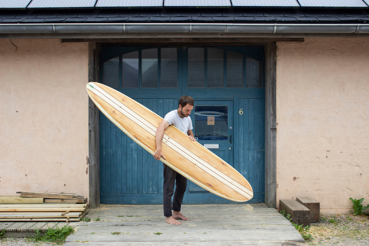 工房のブルーの扉を背景に、長大な中空構造（ホロー）の木製サーフボード（天然木）を抱える裸足のサーファー。クラシカルなロングボードの優美なラインと水平の構図が織りなす、洗練されたエコラグジュアリーなポートレート。