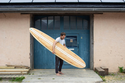 工房のブルーの扉を背景に、長大な中空構造（ホロー）の木製サーフボード（天然木）を抱える裸足のサーファー。クラシカルなロングボードの優美なラインと水平の構図が織りなす、洗練されたエコラグジュアリーなポートレート。