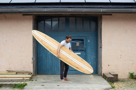 工房のブルーの扉を背景に、長大な中空構造（ホロー）の木製サーフボード（天然木）を抱える裸足のサーファー。クラシカルなロングボードの優美なラインと水平の構図が織りなす、洗練されたエコラグジュアリーなポートレート。
