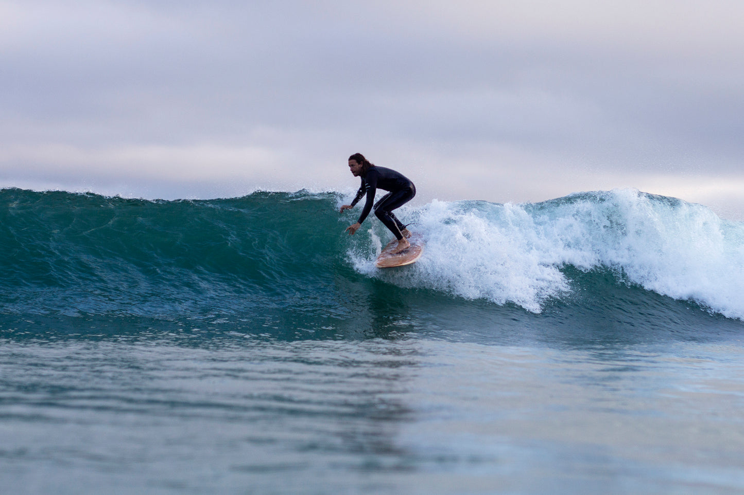 surfer-on-wooden-board-crouched-on-wave A surfer in a black wetsuit, crouched low and trailing a hand in the water, rides the open face of a clean, greenish-blue wave on a hollow wooden surfboard. The wave is breaking behind him with white spray. The sky is overcast, providing soft, diffused light. The photo is taken from the water, capturing the dynamic action.