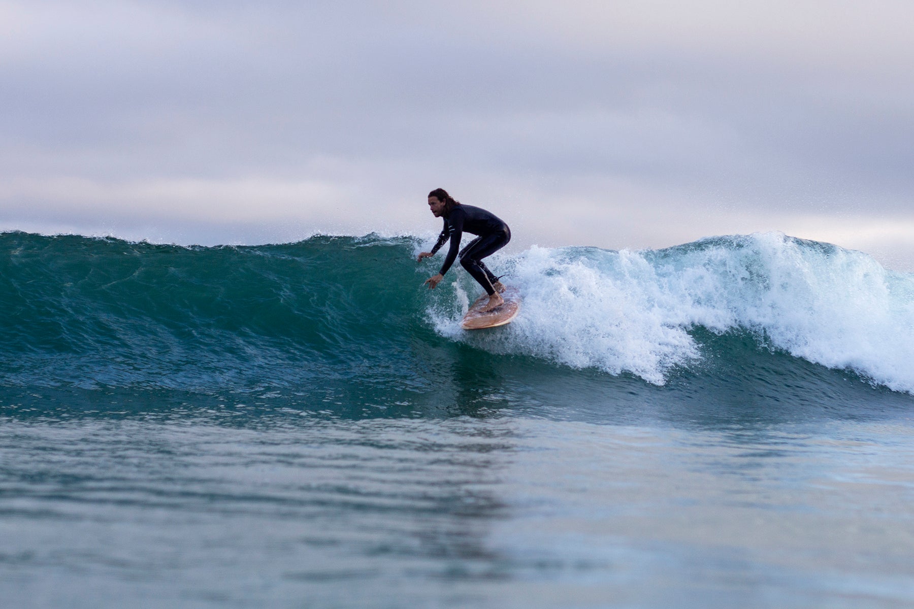 surfer-on-wooden-board-crouched-on-wave A surfer in a black wetsuit, crouched low and trailing a hand in the water, rides the open face of a clean, greenish-blue wave on a hollow wooden surfboard. The wave is breaking behind him with white spray. The sky is overcast, providing soft, diffused light. The photo is taken from the water, capturing the dynamic action.