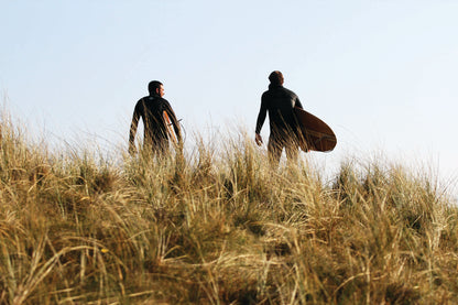 黄金色の草原と、サーファーが抱える中空構造（ホロー）の木製サーフボード（天然木）の色彩の調和。乾いた草のテクスチャーと滑らかなウッドの質感が共鳴する、飾れる・乗れるサステナブルな機能美。