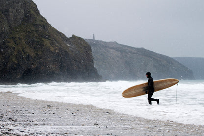 雪が舞う極寒のビーチ、中空構造（ホロー）の木製サーフボード（天然木）を抱えて海へ駆けるサーファー。厳しくも美しい冬の景色に映える温かなウッドの色彩、洗練されたエコラグジュアリーな冒険心。