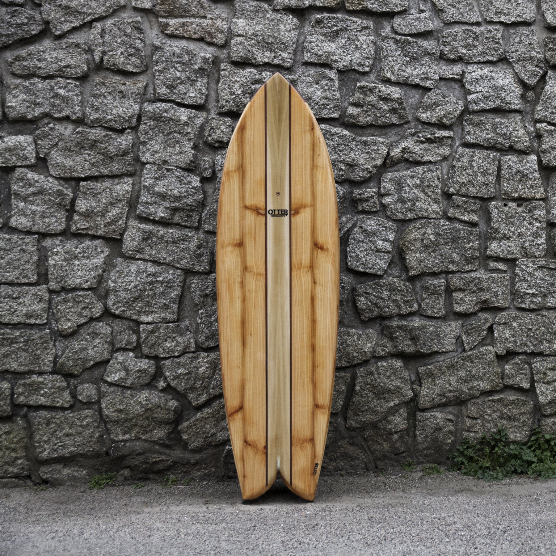 冷たく硬質なグレーの石積みと、温かみのある中空構造（ホロー）の木製サーフボード（天然木）の対比。節（フシ）のある豊かな木の表情と、不揃いな石の凹凸が織りなす、飾れる・乗れるサステナブルな機能美と素材感のコントラスト。