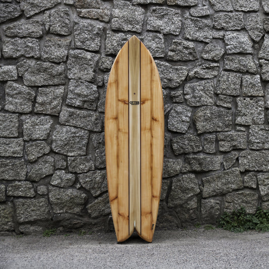 冷たく硬質なグレーの石積みと、温かみのある中空構造（ホロー）の木製サーフボード（天然木）の対比。節（フシ）のある豊かな木の表情と、不揃いな石の凹凸が織りなす、飾れる・乗れるサステナブルな機能美と素材感のコントラスト。